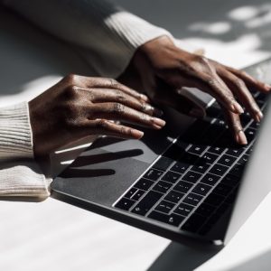 A dark-skinned hand, wearing a white long-sleeve blouse, typing on a silver and black laptop keyboard resting on a table. Accessibility Training & Marketing