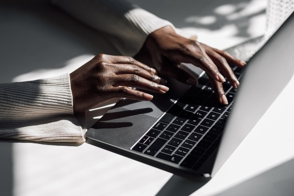 A dark-skinned hand, wearing a white long-sleeve blouse, typing on a silver and black laptop keyboard resting on a table. Accessibility Training & Marketing